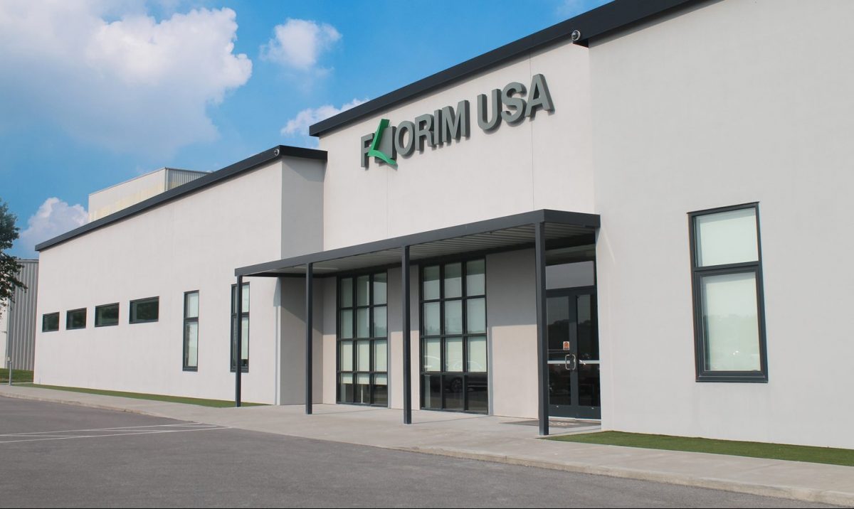 A modern, white industrial building with large windows and a sign reading "Florim USA" above the entrance, under a blue sky with scattered clouds.