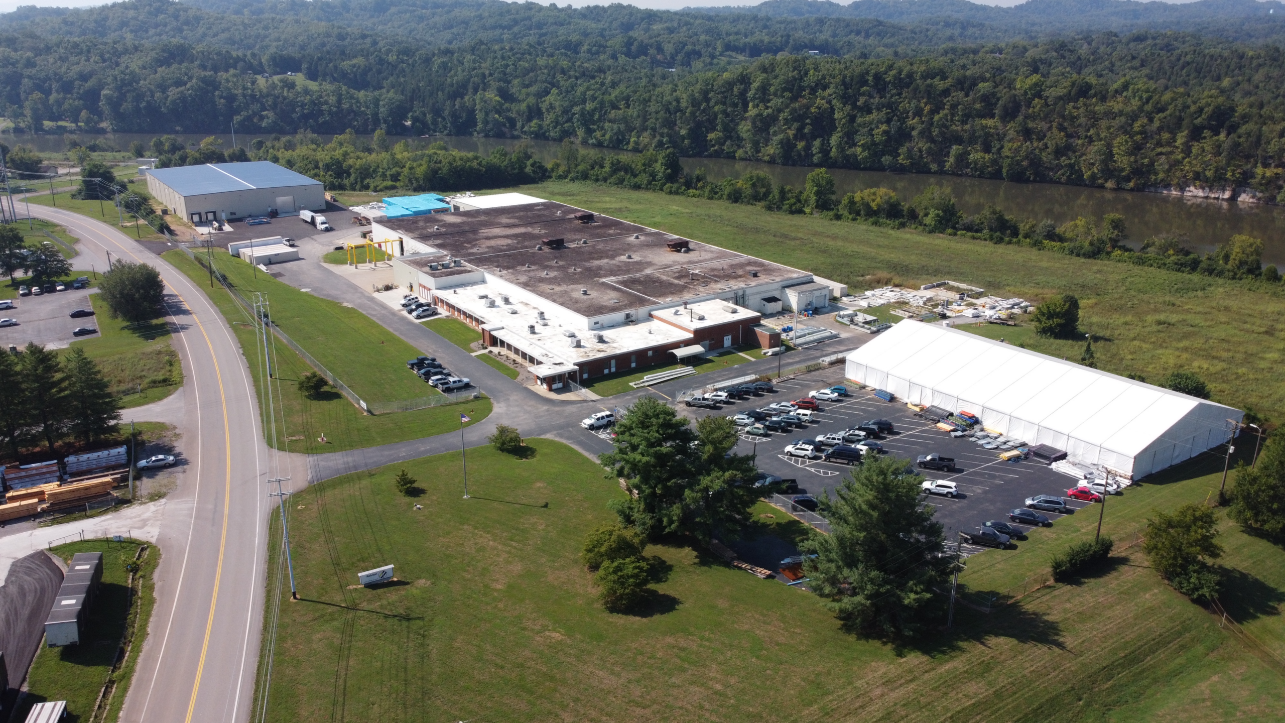 Aerial view of a large industrial facility with white roofs, several parked cars, and a large white tent beside the main building, surrounded by greenery and bordered by a river and trees.