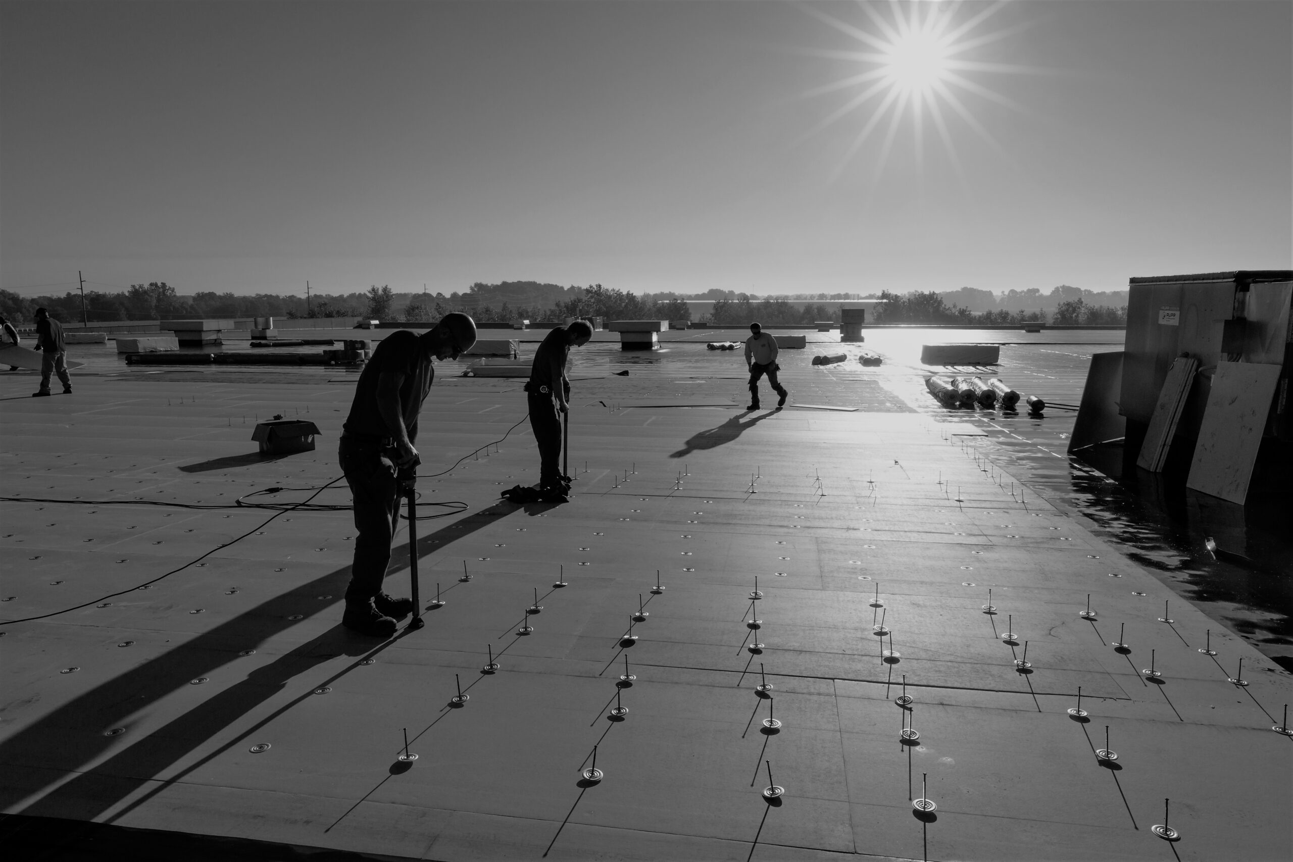 Four people work on a large flat rooftop under a bright sun, securing materials to the roof. Long shadows stretch across the surface, and equipment is scattered nearby. The scene is in black and white.