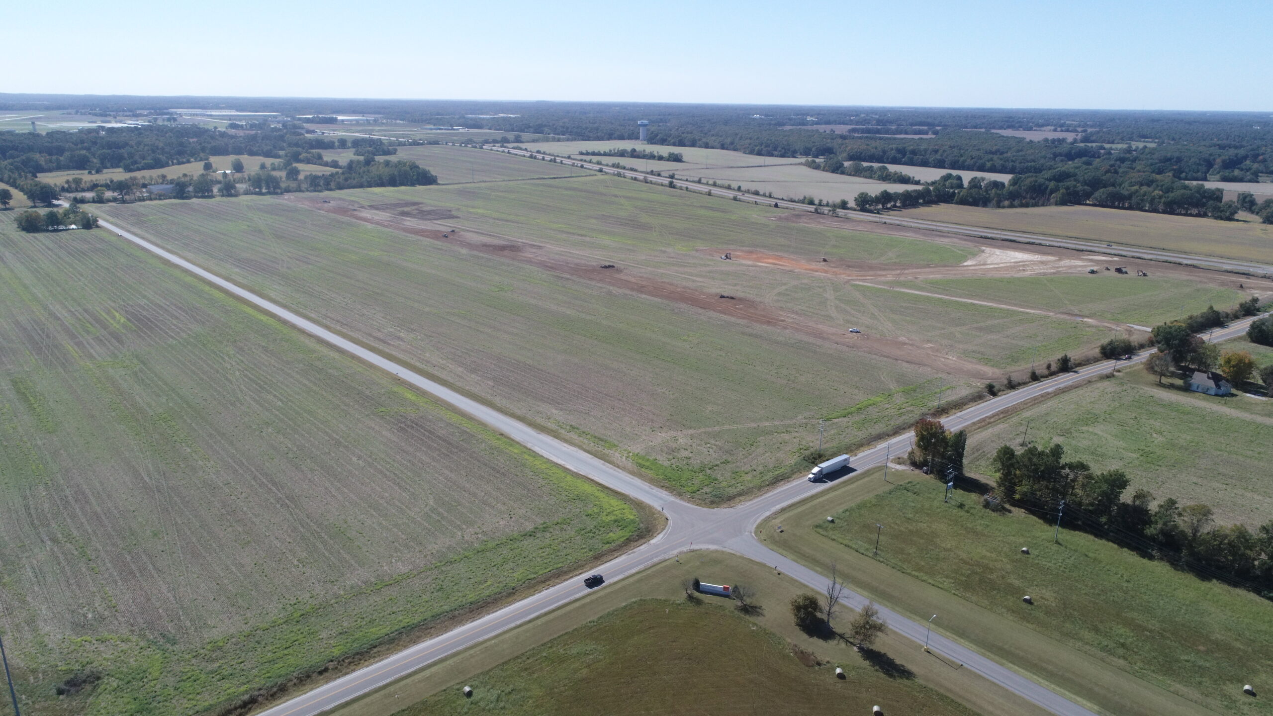 Aerial view of a large, flat, open field intersected by a paved road, with a few vehicles and sparse trees. The land appears mostly undeveloped and stretches toward the horizon under a clear blue sky.