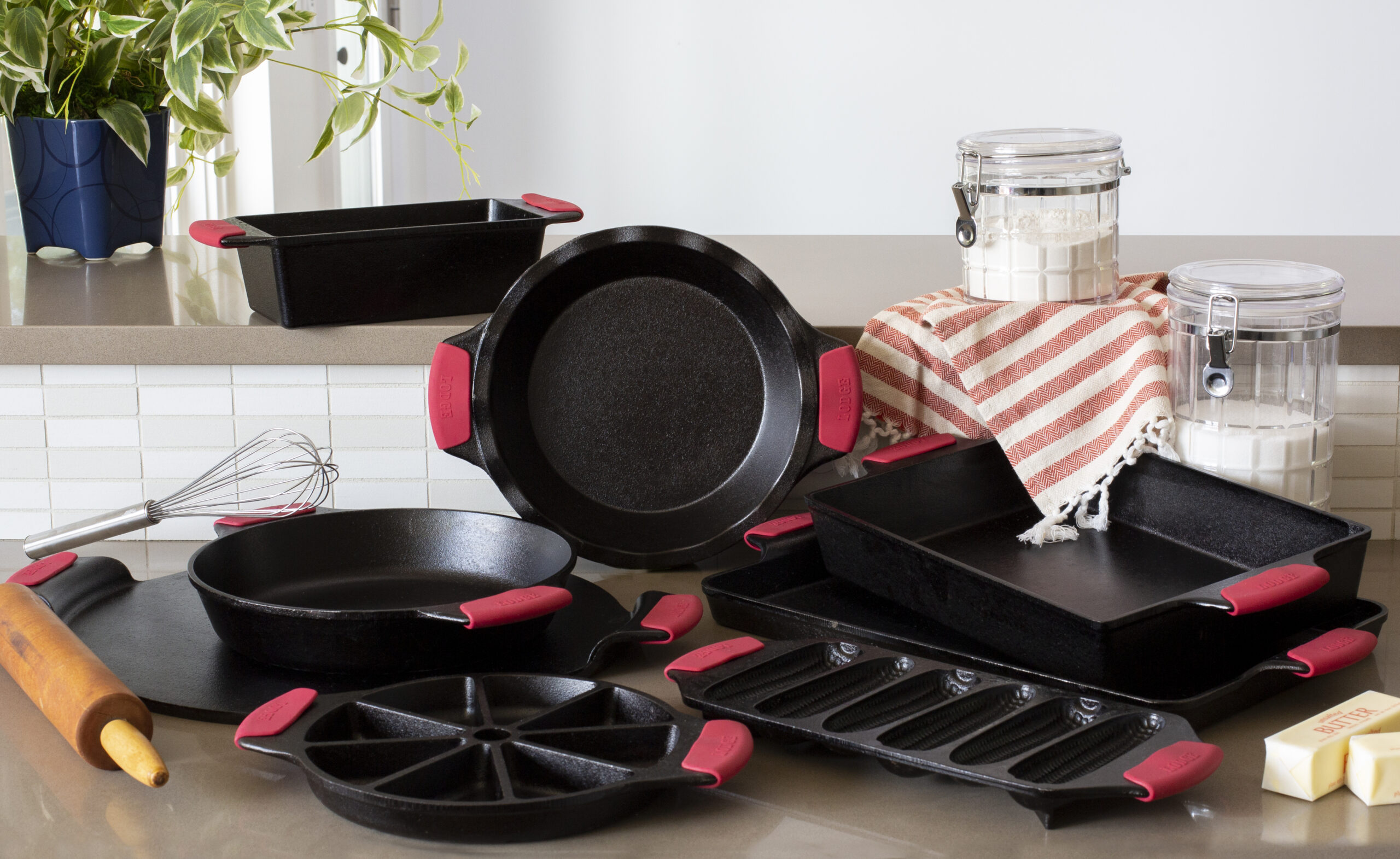 A variety of black cast iron bakeware with red handles is arranged on a kitchen counter with jars of flour and sugar, a whisk, a striped towel, a rolling pin, and a stick of butter nearby.