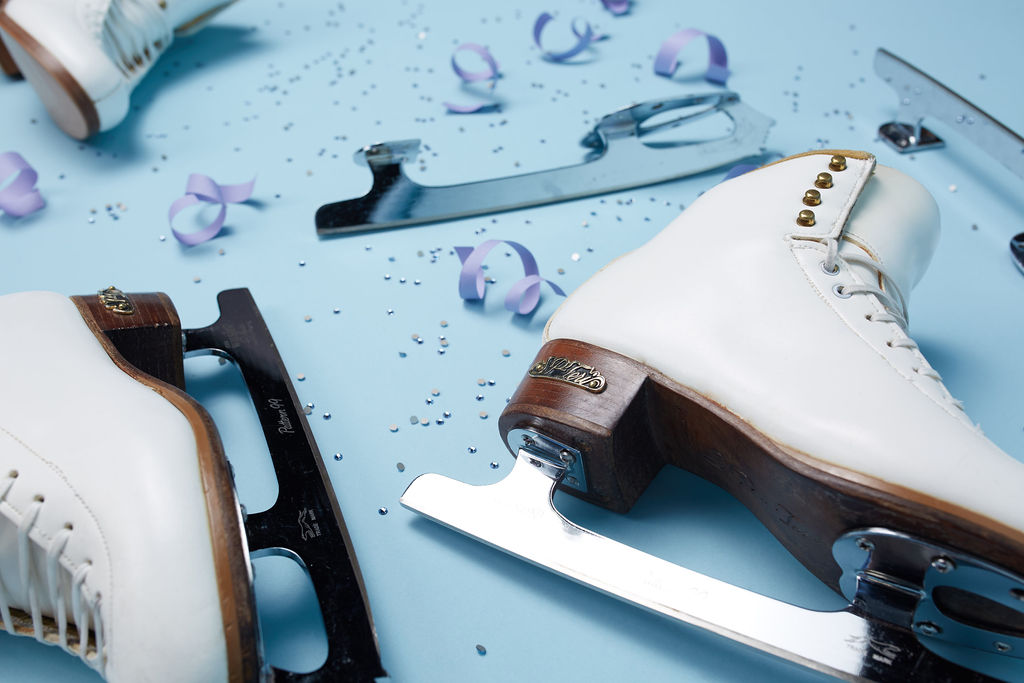 White figure skates with silver blades rest on a light blue surface scattered with purple ribbon curls and small silver confetti.
