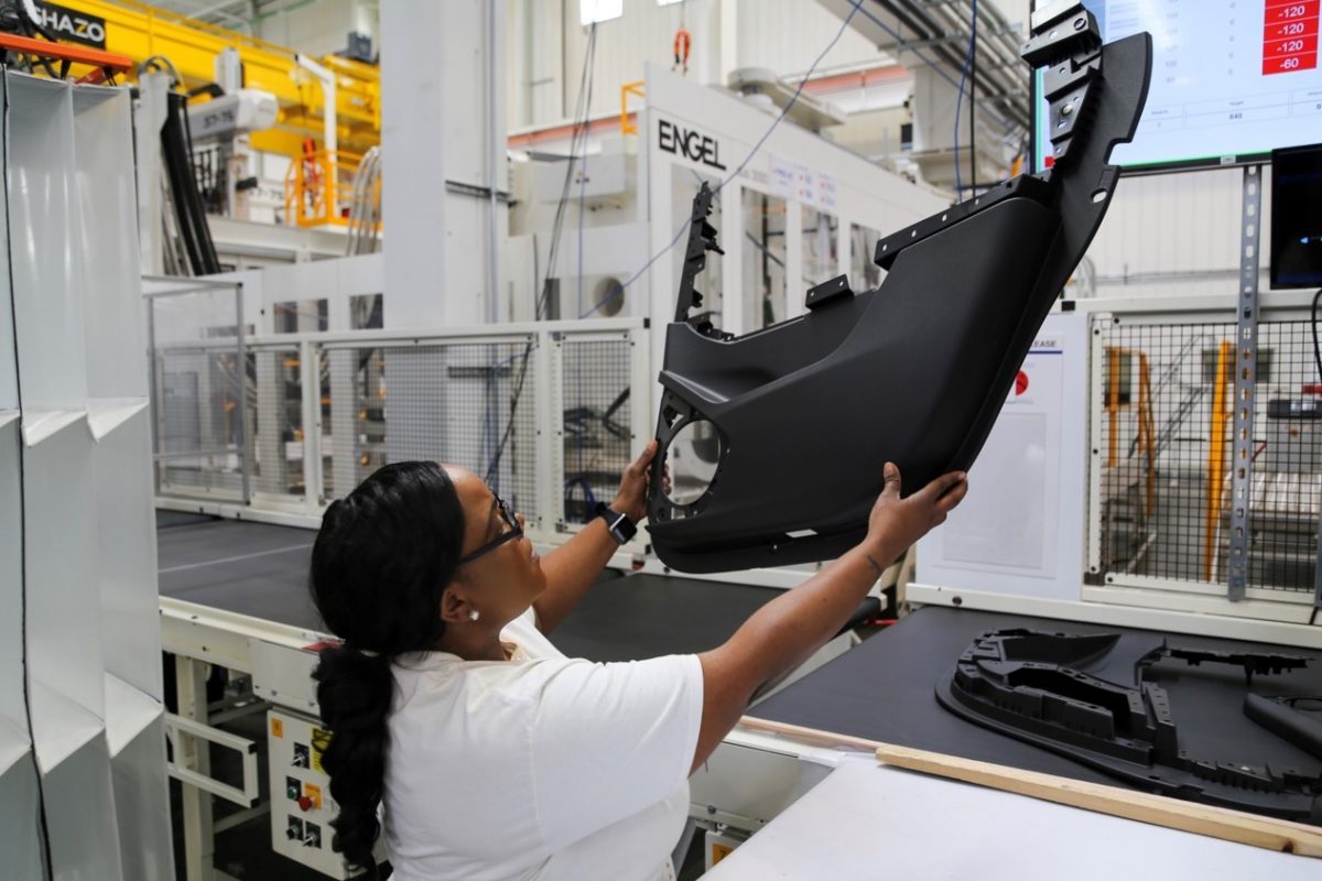 A person wearing glasses and a white shirt inspects a large black automotive part in a factory setting with machinery and screens in the background.