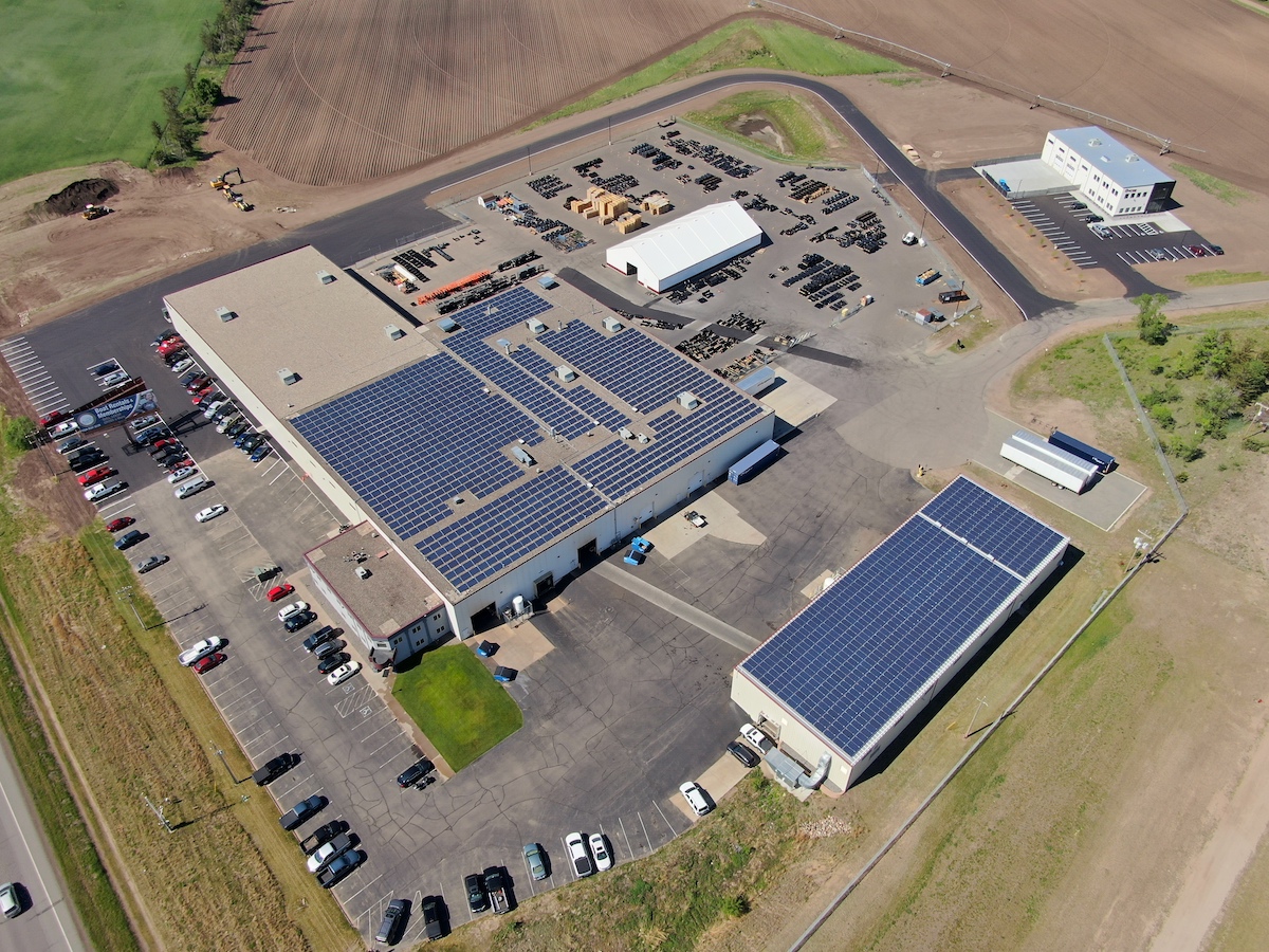 Aerial view of a large industrial complex with solar panels on the roofs, surrounded by parking lots, fields, and several smaller buildings. Numerous cars are parked outside the main structures.