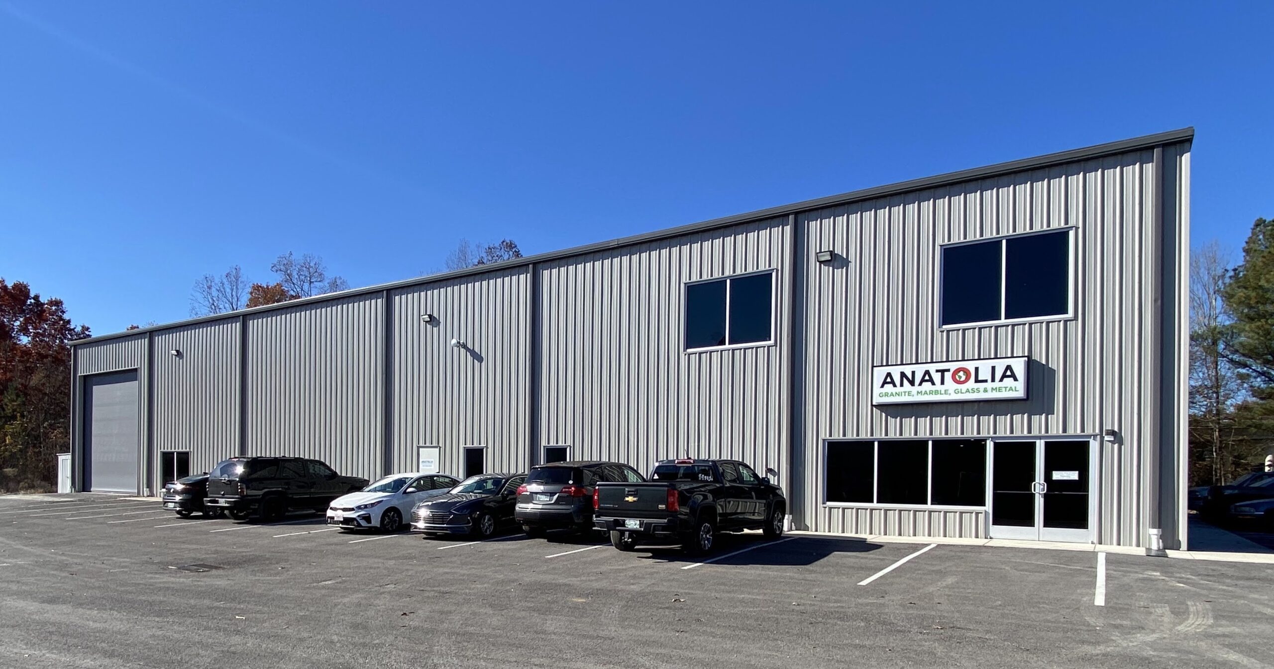 A modern gray metal warehouse building with several windows and parked cars in front. A sign above the entrance reads "ANATOLIA." Clear blue sky and some trees are visible in the background.