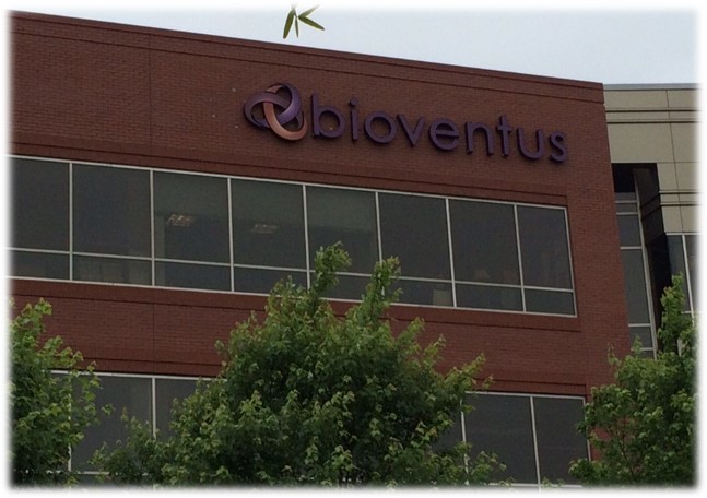 A brick office building with large windows and the "bioventus" logo and name displayed at the top, partially obscured by green leafy trees in the foreground.