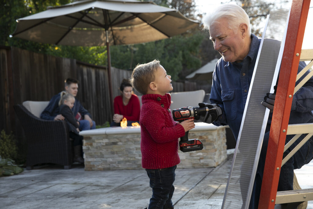 A smiling older man and a young boy hold power tools near a ladder outdoors. Behind them, a family sits by a fire pit on a patio under an umbrella, enjoying a relaxed moment together.