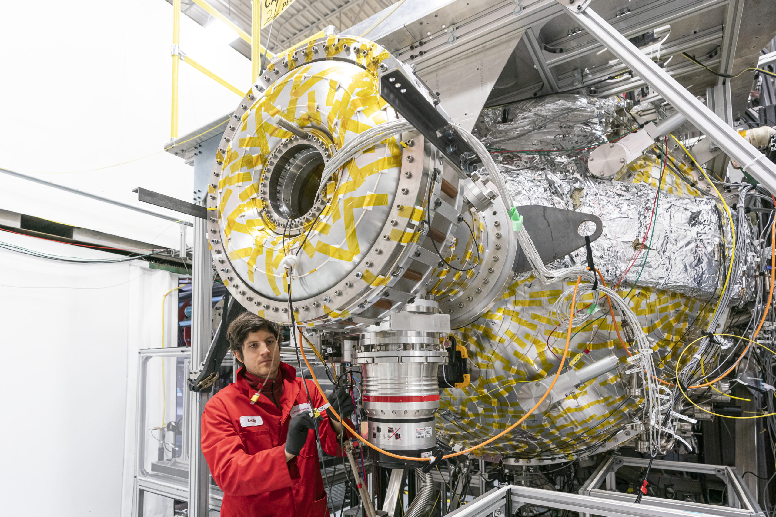 A person in a red lab coat works with a large, cylindrical scientific machine covered in metallic material and yellow tape, surrounded by cables and metal structures in an industrial laboratory.