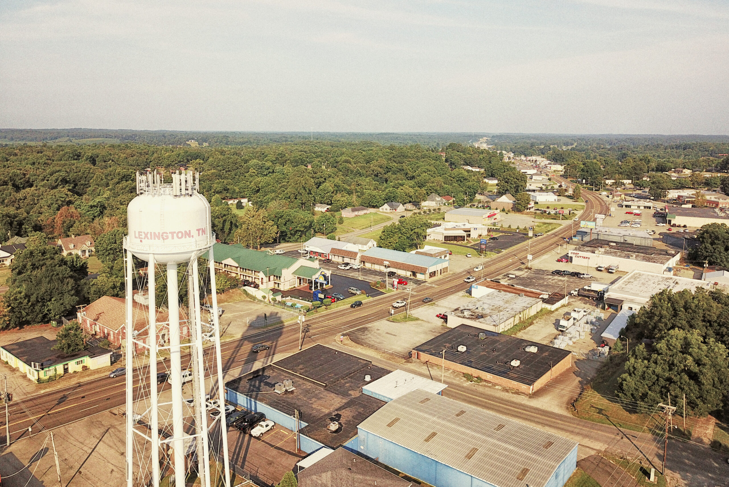 Aerial view of a small town with a white water tower labeled "LEXINGTON, TN," surrounded by buildings, trees, and roads stretching toward the horizon under a clear sky.
