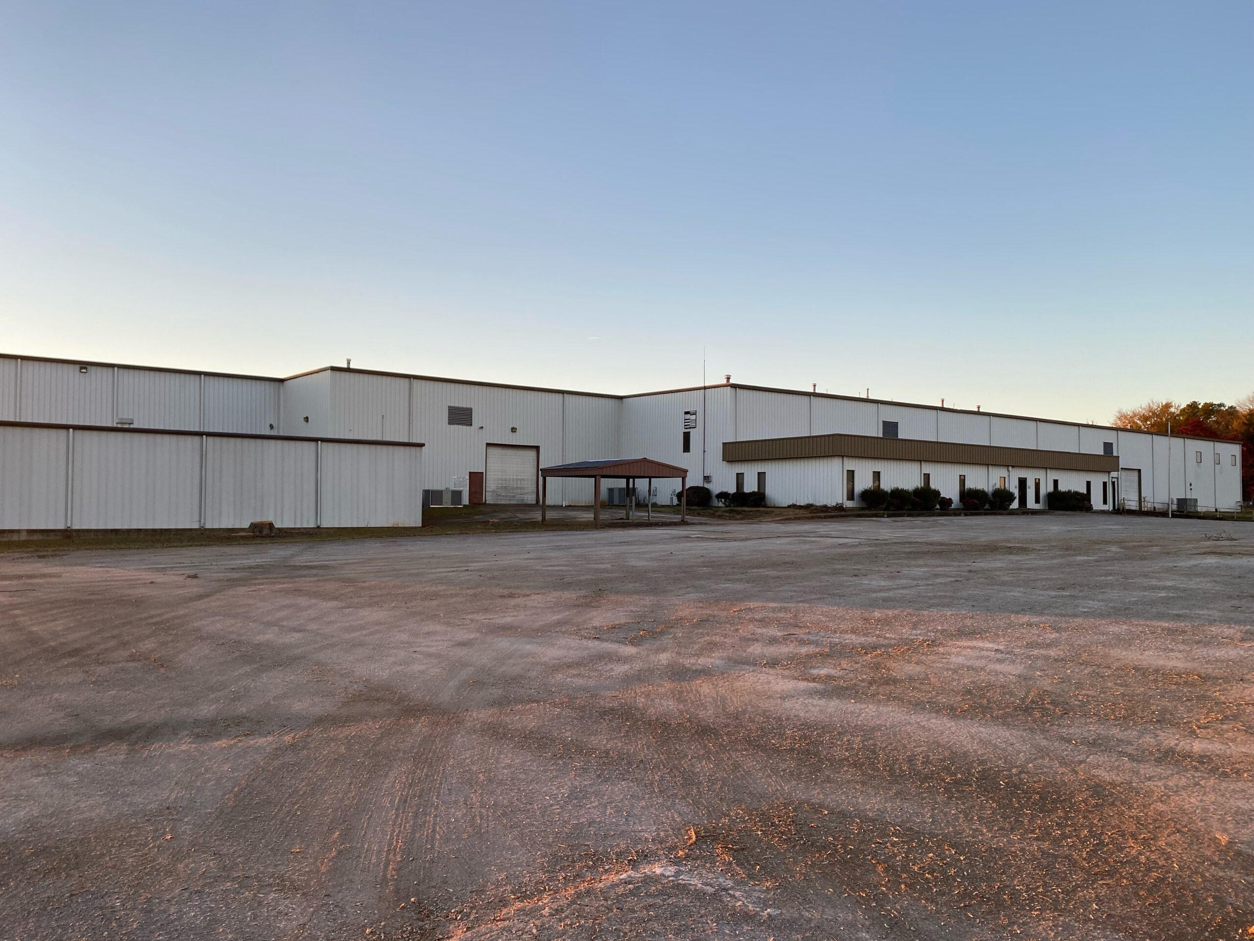 A large, white industrial warehouse building with a flat roof, small windows, and a covered entryway, surrounded by an empty gravel parking lot at sunset.
