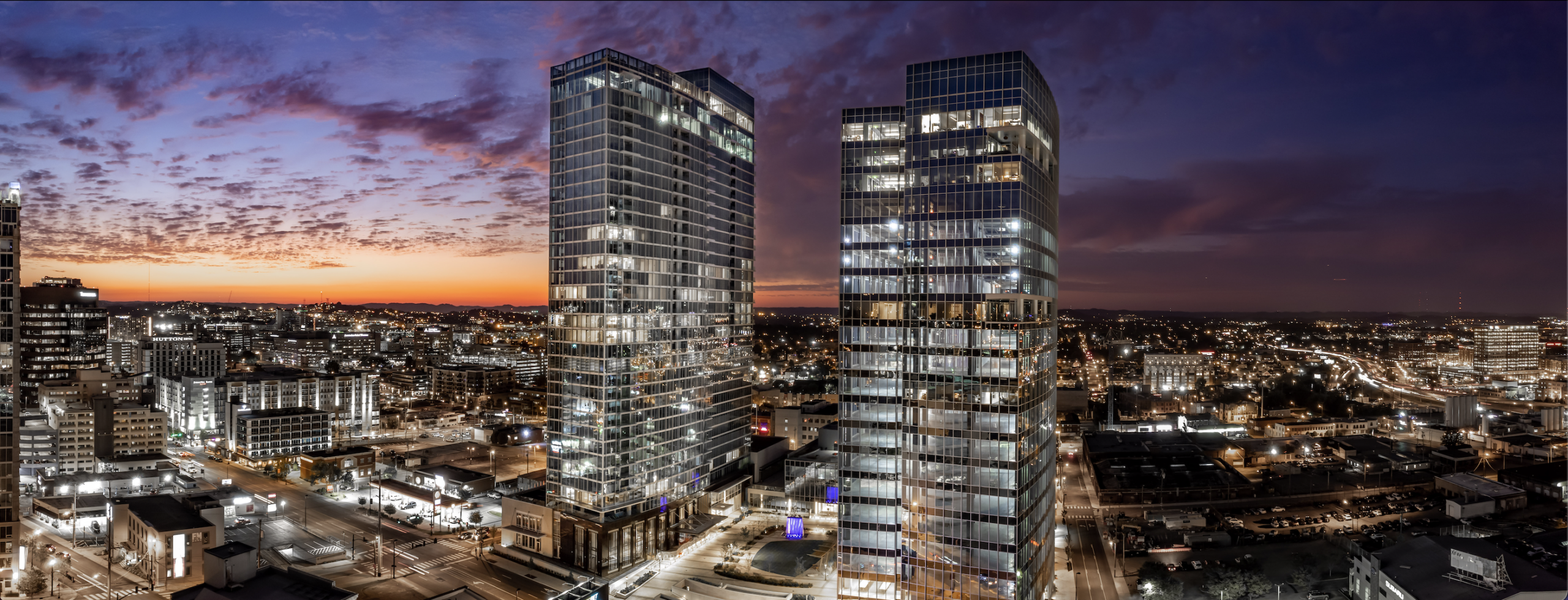 Panoramic view of a cityscape at dusk with two tall, glass office towers reflecting city lights, surrounded by smaller buildings and illuminated streets, under a colorful sunset sky.