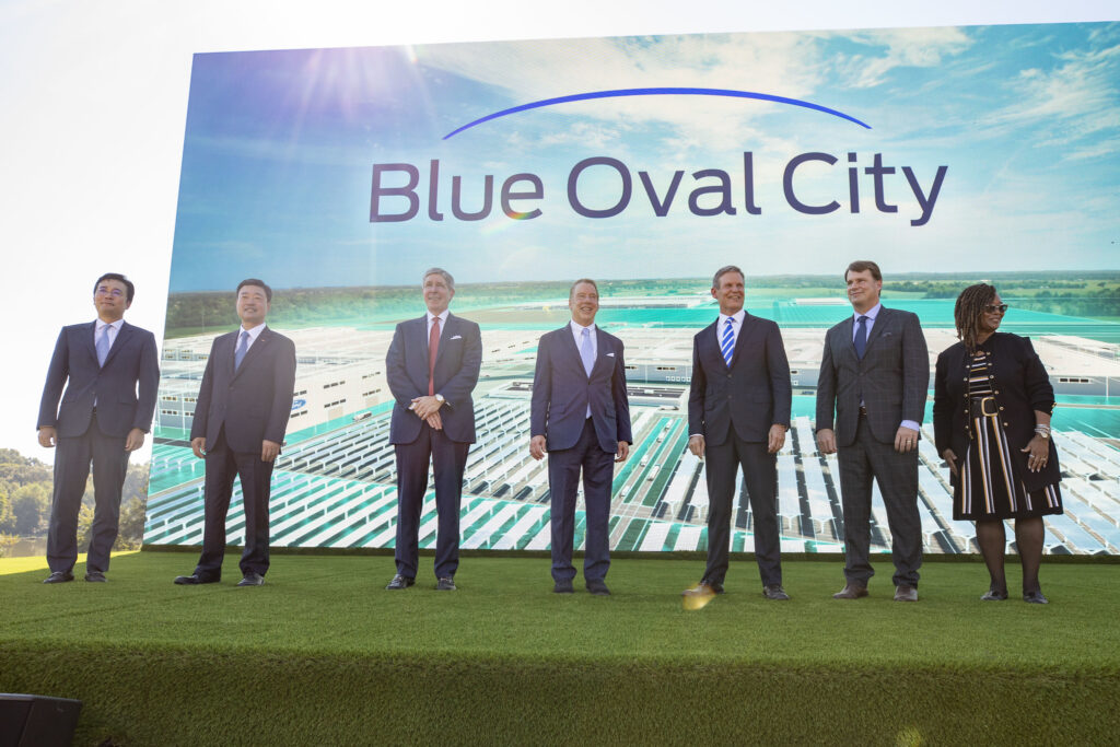 Seven people in business attire stand on a stage with a large screen behind them displaying "Blue Oval City" and an aerial view of an industrial complex. The event appears to be outdoors on a sunny day.