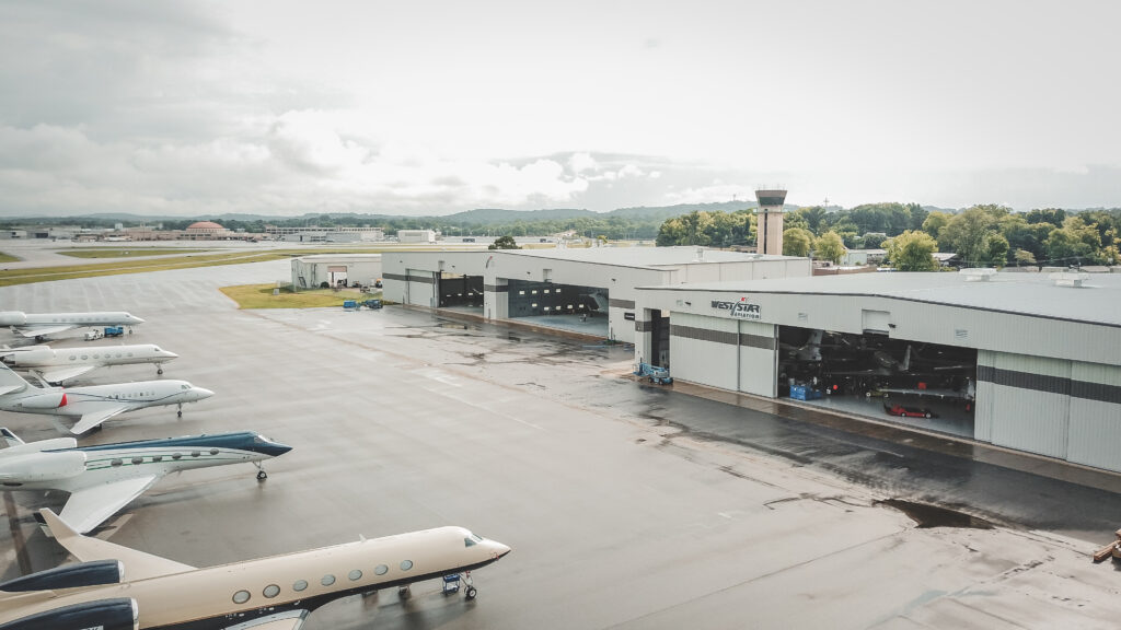 A row of private jets is parked on a wet tarmac near large airport hangars with an air traffic control tower in the background under an overcast sky.