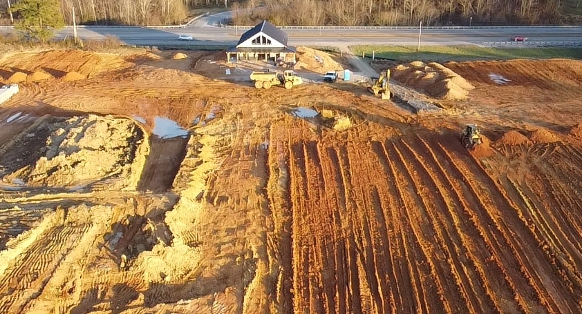 Aerial view of a construction site with reddish soil, heavy machinery, and a small white building near a road. The ground appears to be graded, with visible tire tracks and areas of standing water.