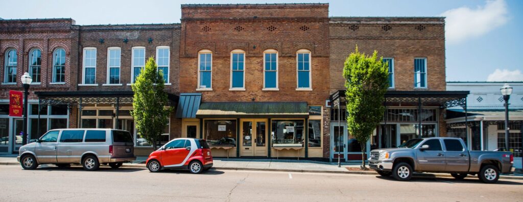 A row of brick storefronts with large windows in a small town, three vehicles parked on the street in front— a silver van, a small red car, and a gray pickup truck— with trees and lampposts along the sidewalk.