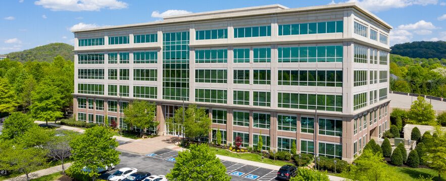 A modern, six-story office building with large glass windows, surrounded by green trees and landscaping, with a parking lot featuring accessible parking spaces in the foreground under a partly cloudy sky.