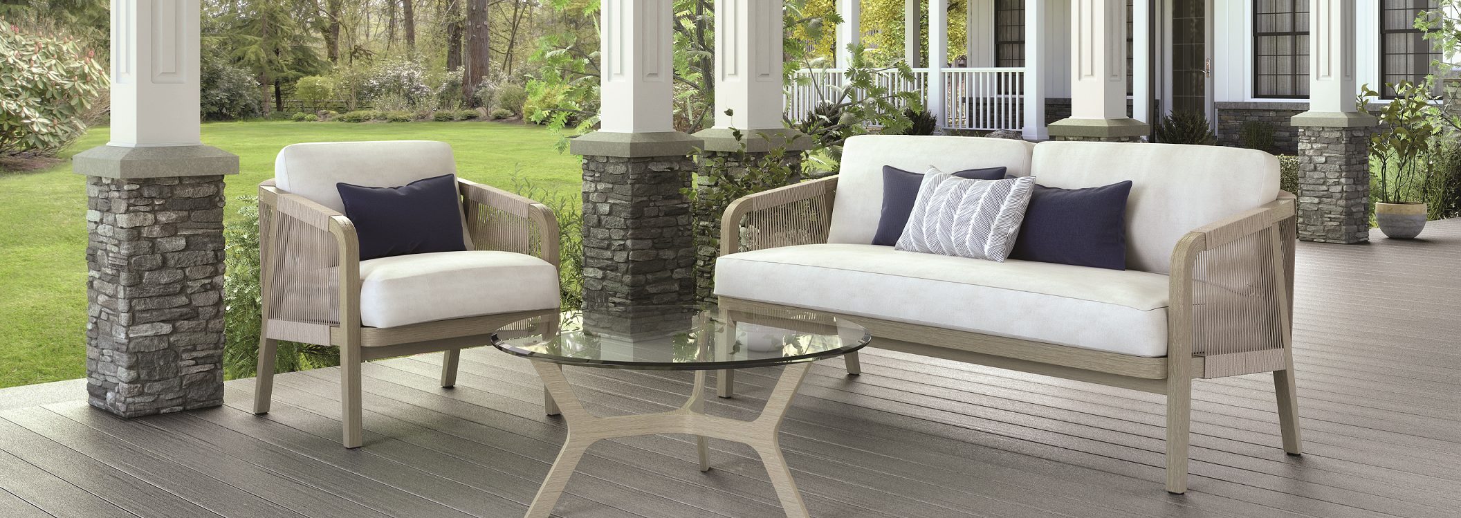 Outdoor patio with a cushioned armchair and sofa, both with throw pillows, and a round glass coffee table. The area features stone pillars and overlooks a green lawn and trees.