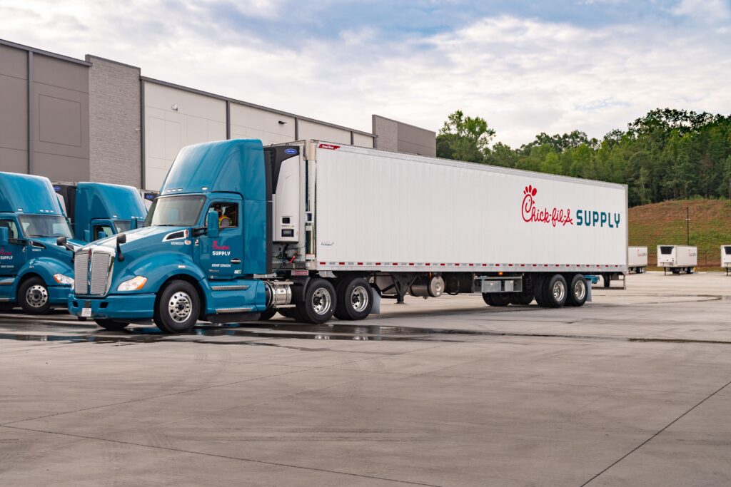 A blue semi-truck with a white trailer labeled "Chick-fil-A Supply" is parked in an industrial lot near a warehouse, with other similar trucks and trees in the background.