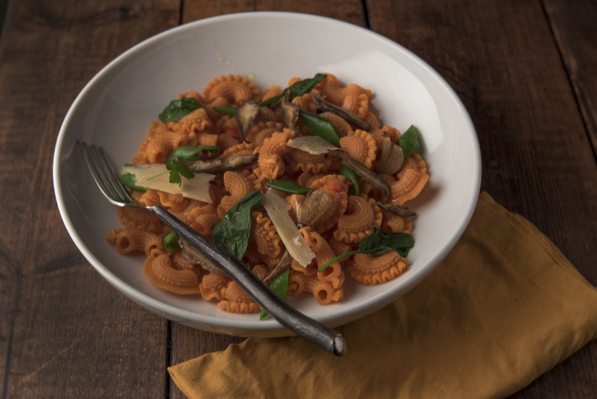 A white bowl of pasta with tomato sauce, sautéed mushrooms, spinach, and shaved parmesan sits on a wooden table with a fork and a mustard yellow napkin beside it.