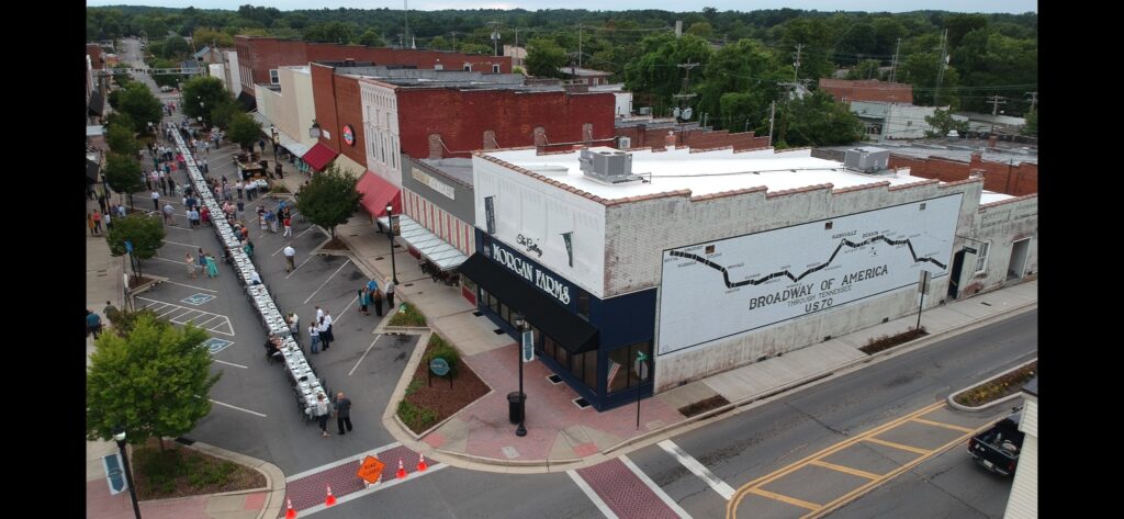 Aerial view of a small town street with people gathered around a long table set up for an outdoor event. The street is lined with brick buildings, one displaying a mural that says "Broadway of America, 1917-1927.