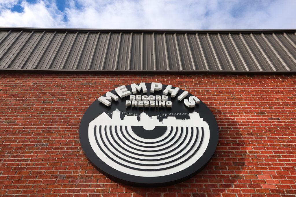 A large circular sign on a brick wall reads "Memphis Record Pressing," featuring the Memphis Record Pressing city skyline and a vinyl record design, set against a blue sky with clouds in the background.