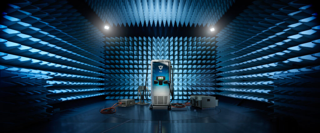 A tall charging station stands in the center of an anechoic chamber with blue, spiky foam walls. Electronic equipment and cables are on the floor, and overhead lights illuminate the futuristic scene.