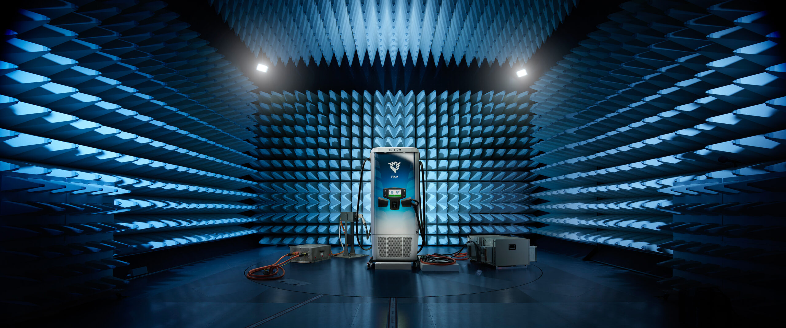 A tall charging station stands in the center of an anechoic chamber with blue, spiky foam walls. Electronic equipment and cables are on the floor, and overhead lights illuminate the futuristic scene.