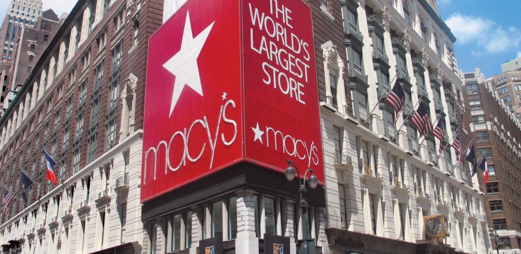 A large red sign on a corner building reads "The World's Largest Store, Macy's" with a white star, on the exterior of Macy's department store in New York City. The building has American flags and decorative architecture.