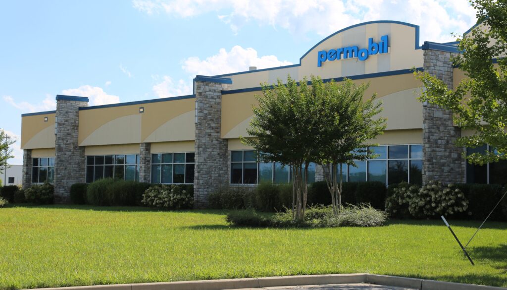 A beige and stone commercial building with the sign "permobil" above the entrance, surrounded by green grass, trimmed bushes, and a few trees under a blue sky with scattered clouds.