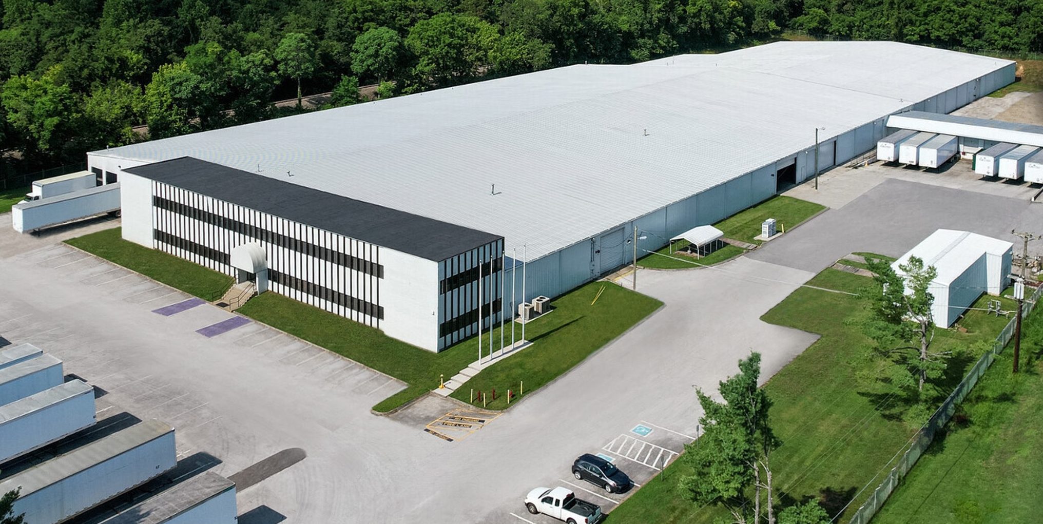 A large white industrial warehouse with a black roof section is surrounded by trucks, loading docks, parking spaces, and green trees in the background.