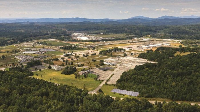 Aerial view of a large industrial complex surrounded by green forests and rolling hills, with mountains visible in the background under a partly cloudy sky.