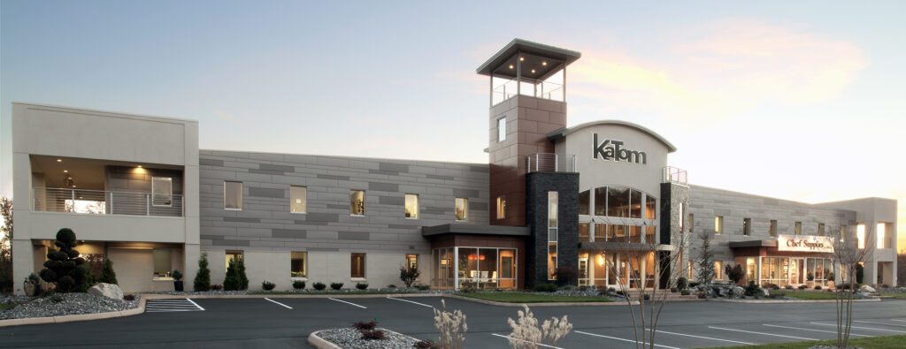 Modern two-story commercial building with “KaTom” signage, large windows, a stone accent wall, and a prominent tower. The parking lot in front is mostly empty, and landscaping includes rocks and small bushes.