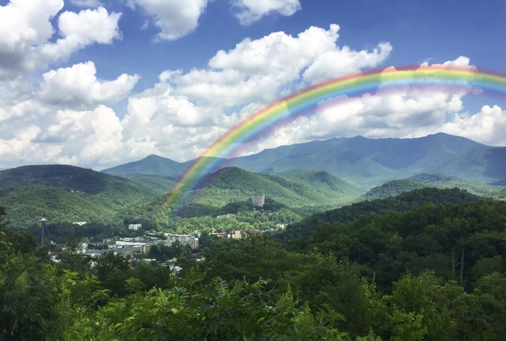 A vivid rainbow arches over a lush, green valley with distant mountains, scattered buildings, and a partly cloudy blue sky.