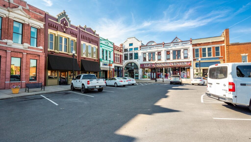 A small-town square with historic, colorful storefronts, parked white vehicles, and clear blue sky above. Store signs are visible, and sunlight casts shadows on the street.