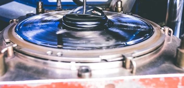 Close-up of a vinyl record being pressed in a record manufacturing machine, with the round disc held in place by metal clamps and a central spindle above it.