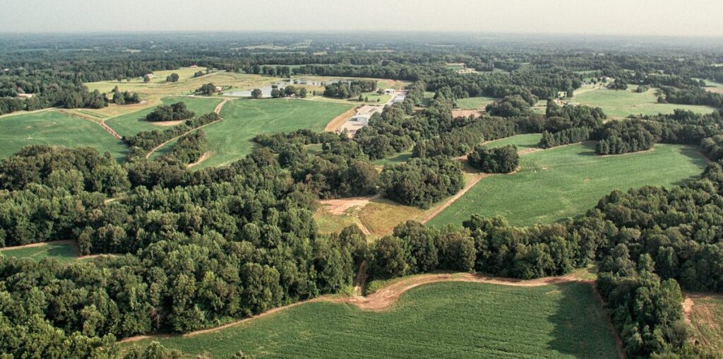 Aerial view of a rural landscape with large green fields, clustered trees, and winding dirt paths under a hazy sky. Buildings and small ponds are visible in the distance among the greenery.