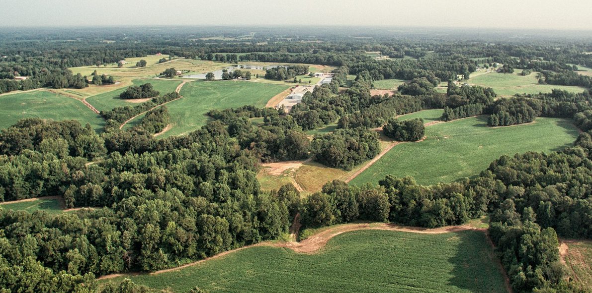 Aerial view of a rural landscape with large green fields, clustered trees, and winding dirt paths under a hazy sky. Buildings and small ponds are visible in the distance among the greenery.