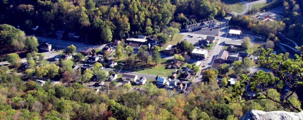 Aerial view of a small town surrounded by dense, green trees, with houses, buildings, streets, and parking lots visible among the foliage.