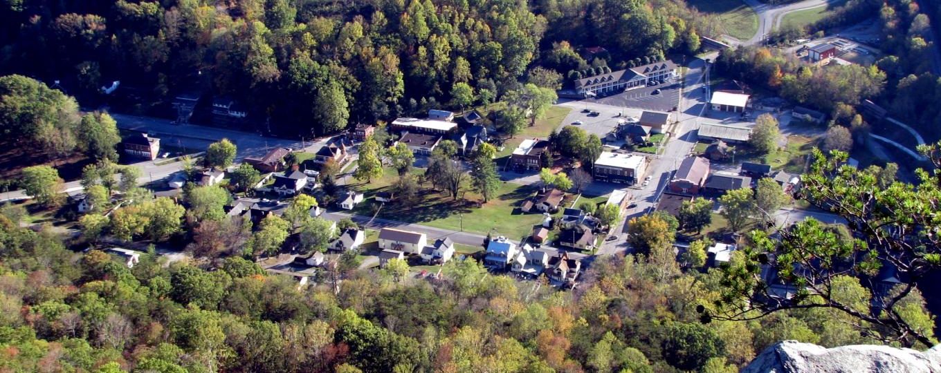 Aerial view of a small town surrounded by dense, green trees, with houses, buildings, streets, and parking lots visible among the foliage.