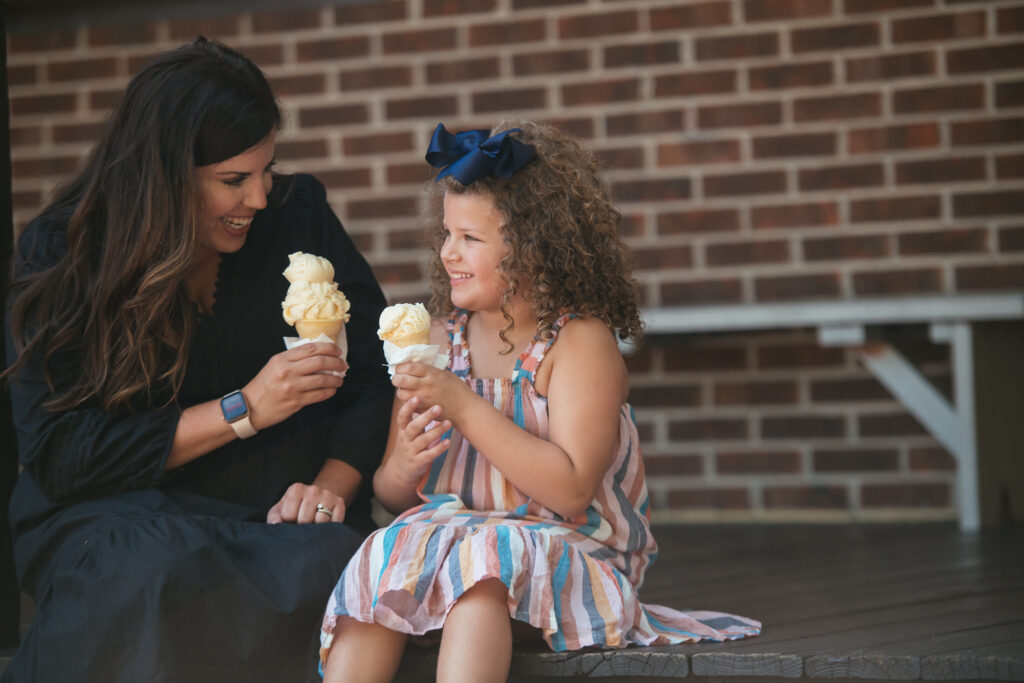 A woman and a young girl sit on a wooden bench, smiling and holding ice cream cones. The girl wears a striped dress and blue bow, while the woman wears a black dress. A brick wall is in the background.