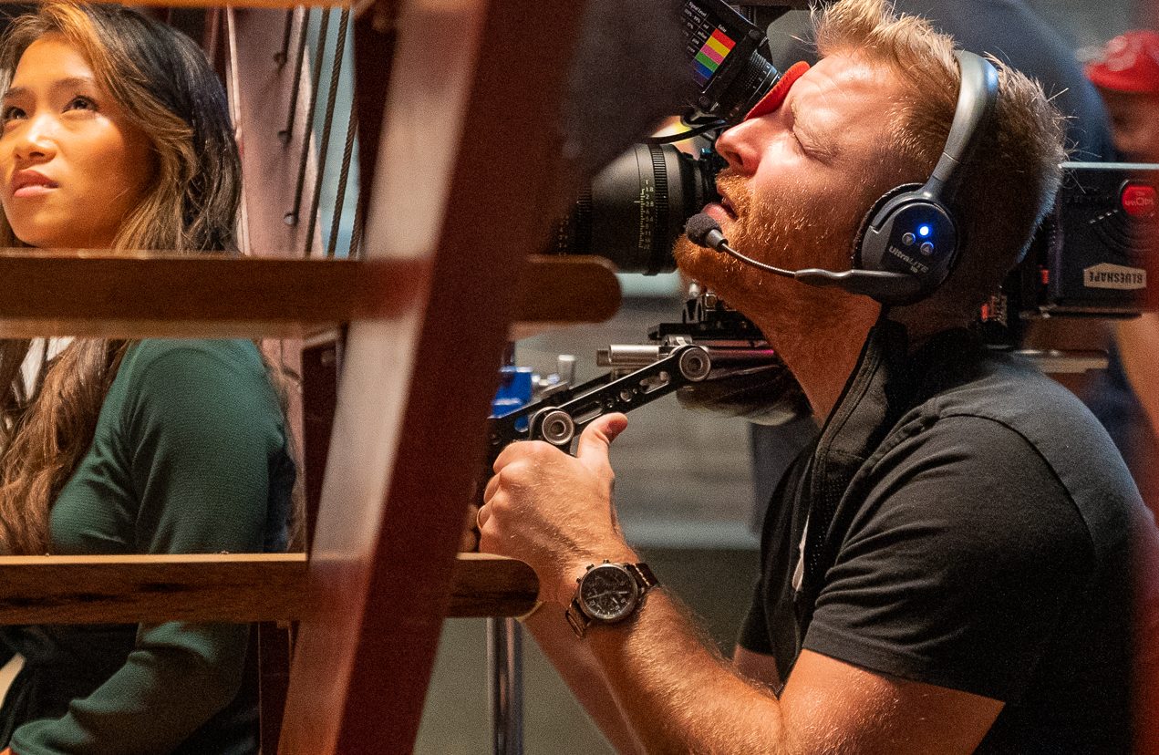 A man with headphones operates a large camera, looking through the viewfinder and focusing intently. Next to him, a woman with long hair in a green top gazes upward. Wooden rails are visible in the foreground.