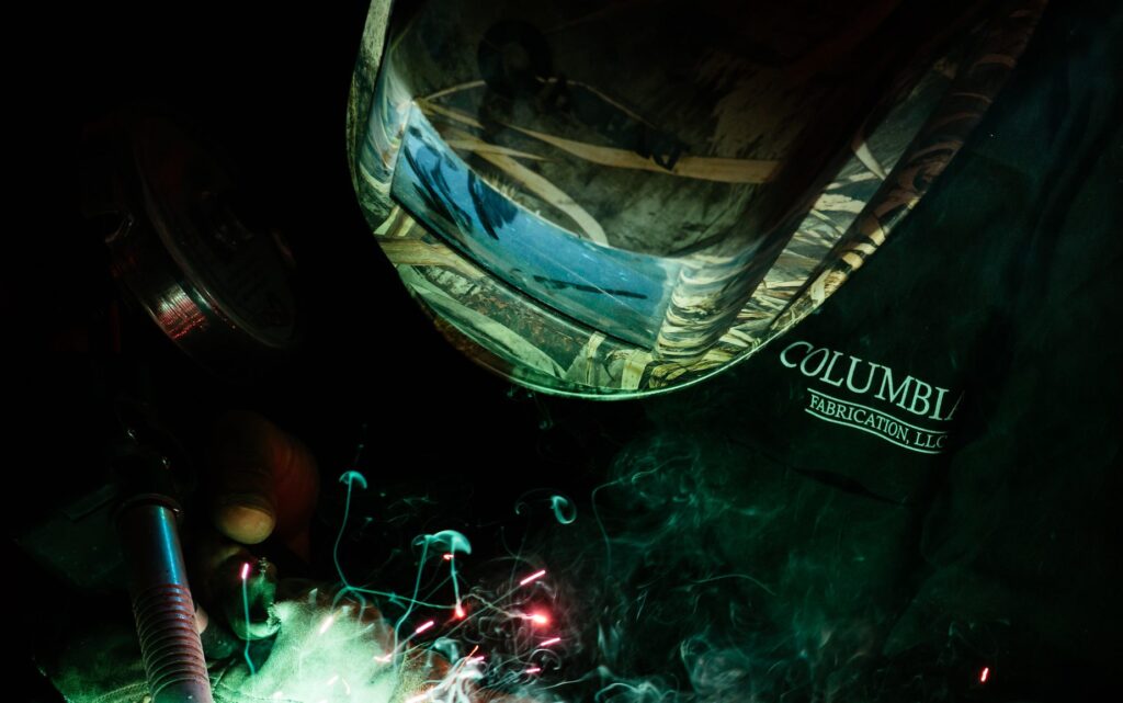 A welder in a camouflage-patterned helmet and a dark jacket labeled "COLUMBI FABRICATION, LLC" works with bright sparks and smoke rising in a dimly lit environment.