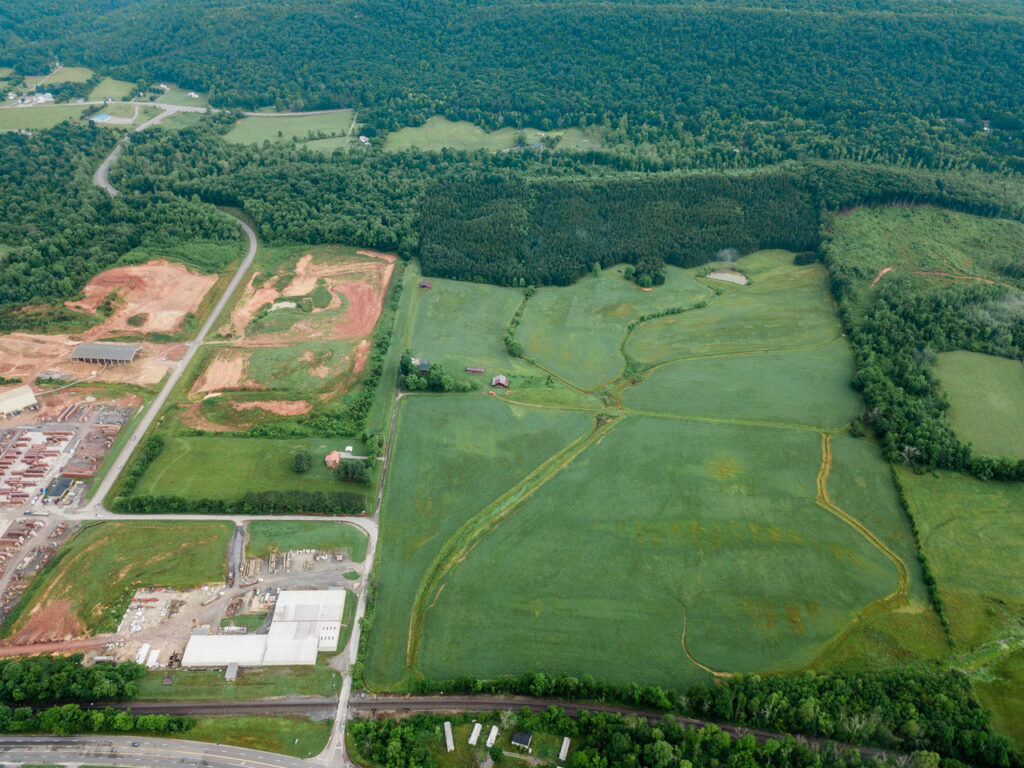 Aerial view of a rural landscape showing green fields, scattered trees, forested hills, and some industrial buildings along roads. The area includes patches of cleared land and several farm structures.