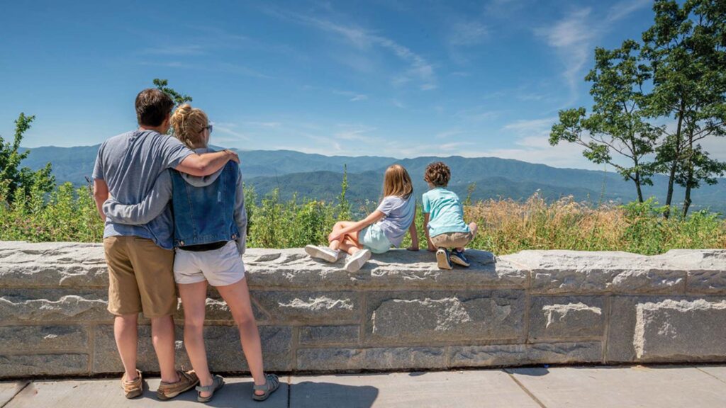 A couple stands arm in arm while two children sit on a stone wall, all looking out at a scenic view of mountains and blue sky on a sunny day.