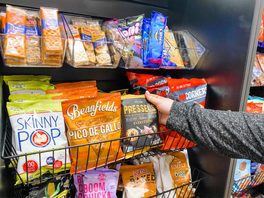 A person’s hand reaches for a bag of PopCorners chips on a snack shelf filled with various packaged snacks, including Beanfields chips, Skinny Pop popcorn, Pretzel Crisps, and cookies.