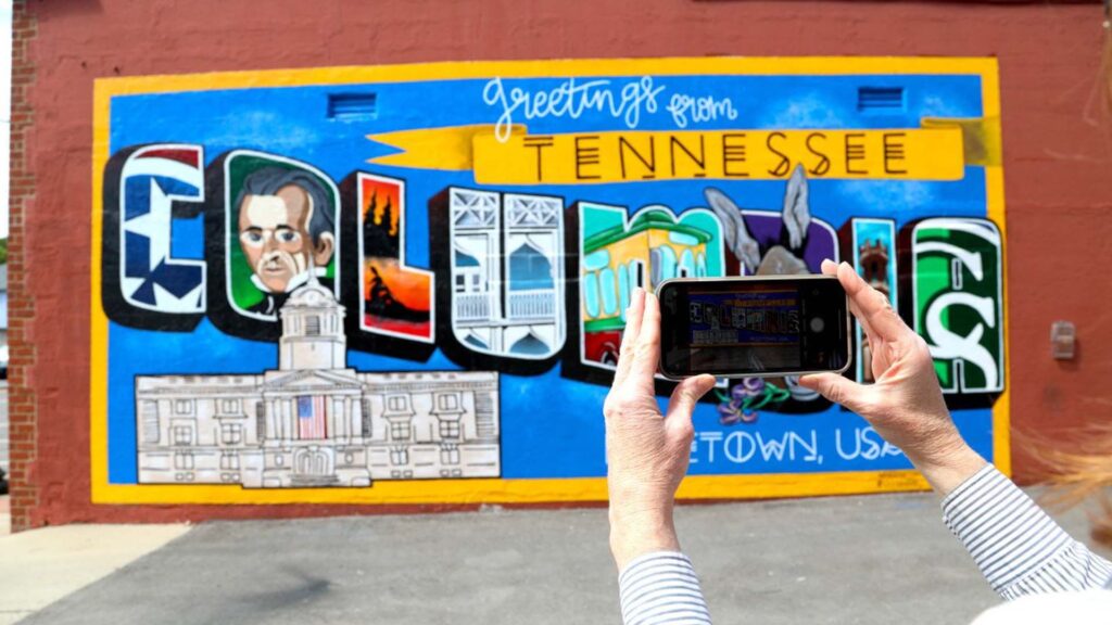 A person holds up a phone to photograph a colorful mural that says "Greetings from Tennessee Columbia" with local landmarks and images painted on a red brick wall.