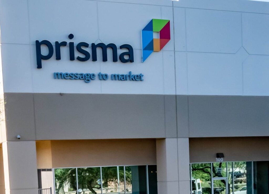 Sign on a building reads "prisma message to market" with a colorful cube logo beside it; the building has beige and white walls and large reflective windows showing trees outside.