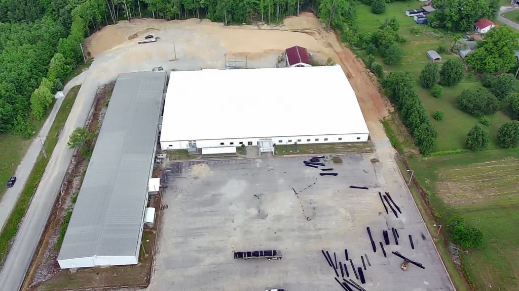 Aerial view of a large, white-roofed industrial building with an adjacent gray-roofed structure, surrounded by empty paved areas, construction materials, and green trees on all sides.