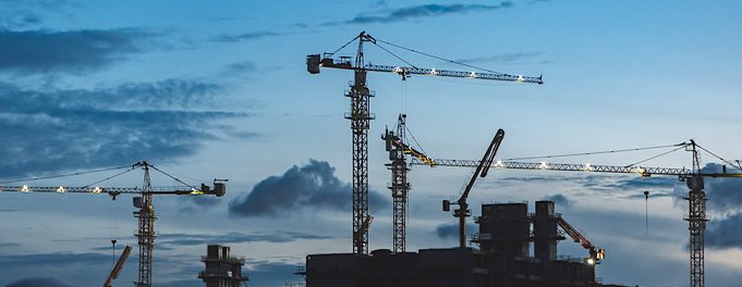 Several construction cranes stand over a building site at dusk, silhouetted against a blue sky with scattered clouds. The cranes' booms stretch across the scene, indicating ongoing construction work.