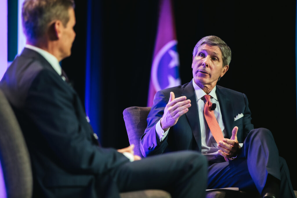 Two men in suits sit and talk on stage, one gesturing with his hand while speaking. A flag with a circular emblem is visible in the background. The setting appears formal, likely a conference or panel discussion.
